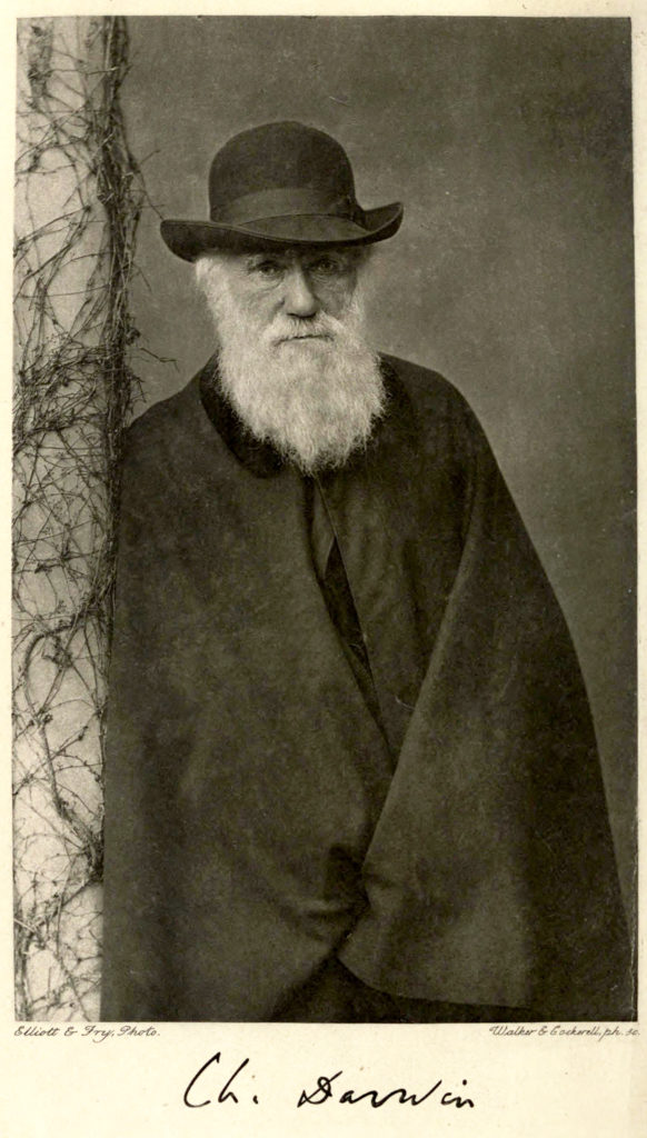 Black and white photo of Charles Darwin leaning against a column, wearing a hat.